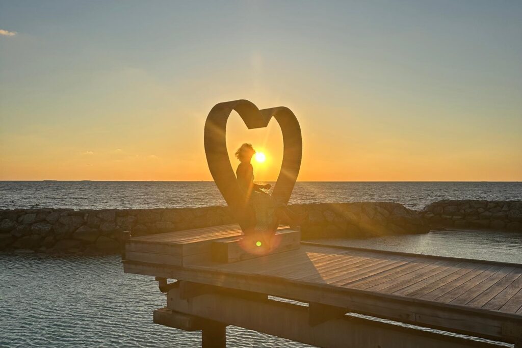 Herz am Strand bei Sonnenuntergang, in dessen Mitte eine sitzende Person silhouettenhaft zu sehen ist.