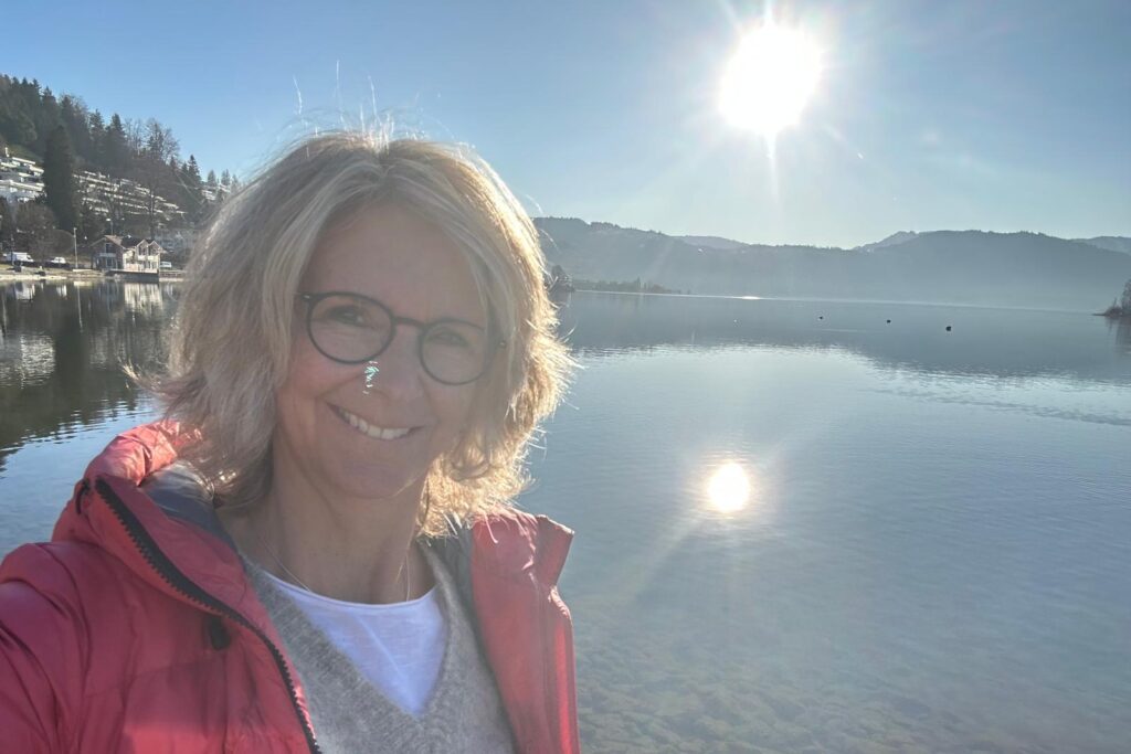 Tanja Baumann lächelt sanft, umgeben von See und Sonnenlicht, eine Stimmung von Ruhe und Herzverbundenheit.